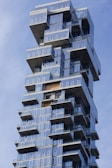 A modern skyscraper featuring a unique design with staggered balconies and large glass windows, creating a visually striking geometric pattern. The building is set against a clear blue sky, enhancing its imposing and innovative architectural style.