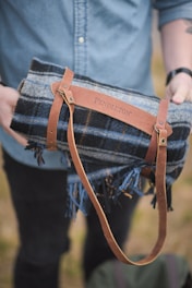 A person holds a rolled-up plaid blanket with a brown leather strap embossed with the word 'Pendleton'. The person is wearing a denim shirt, and the background is a blurred outdoor setting.