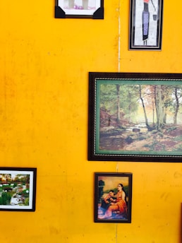 Various framed artworks and photographs hang on a vibrant yellow wall. The artworks include a painting of a serene landscape with trees, a small garden scene, and a classical depiction of a seated woman with a child.