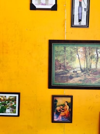 Various framed artworks and photographs hang on a vibrant yellow wall. The artworks include a painting of a serene landscape with trees, a small garden scene, and a classical depiction of a seated woman with a child.
