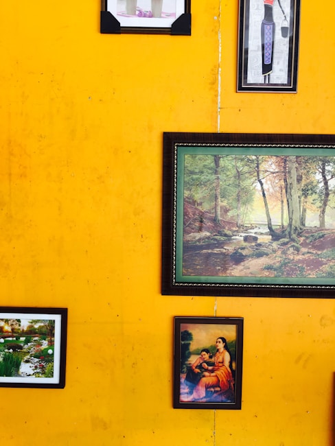 Various framed artworks and photographs hang on a vibrant yellow wall. The artworks include a painting of a serene landscape with trees, a small garden scene, and a classical depiction of a seated woman with a child.
