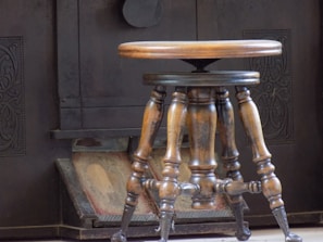 A wooden stool with ornate, turned legs and a round seat stands in front of an old-fashioned, dark metal stove. The stove has intricate carved patterns on its surface, suggesting a vintage design.