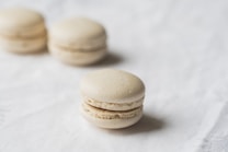 Three light-colored macarons are placed on a soft white surface. The macarons have a smooth and slightly glossy finish, with a creamy filling visible between the top and bottom halves.