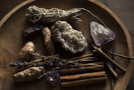 A wooden bowl contains various natural items including a bundle of sage, a cluster of amethyst, a large raw crystal, dried lavender sprigs, ginger roots, and cinnamon sticks. The setting has a rustic and earthy feel, with dim lighting enhancing the natural textures and colors of the objects.