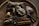 A wooden bowl contains various natural items including a bundle of sage, a cluster of amethyst, a large raw crystal, dried lavender sprigs, ginger roots, and cinnamon sticks. The setting has a rustic and earthy feel, with dim lighting enhancing the natural textures and colors of the objects.