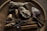 A wooden bowl contains various natural items including a bundle of sage, a cluster of amethyst, a large raw crystal, dried lavender sprigs, ginger roots, and cinnamon sticks. The setting has a rustic and earthy feel, with dim lighting enhancing the natural textures and colors of the objects.