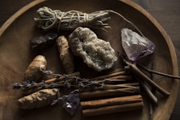 A wooden bowl contains various natural items including a bundle of sage, a cluster of amethyst, a large raw crystal, dried lavender sprigs, ginger roots, and cinnamon sticks. The setting has a rustic and earthy feel, with dim lighting enhancing the natural textures and colors of the objects.