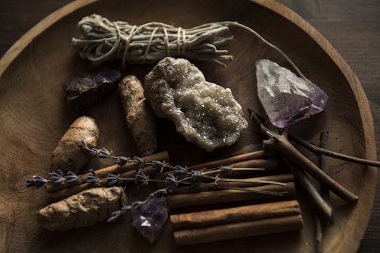 A wooden bowl contains various natural items including a bundle of sage, a cluster of amethyst, a large raw crystal, dried lavender sprigs, ginger roots, and cinnamon sticks. The setting has a rustic and earthy feel, with dim lighting enhancing the natural textures and colors of the objects.