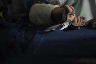 A hand holding a pair of metallic scissors is visible, poised to cut through a piece of dark fabric. The setting appears to be dimly lit, suggesting a focused, intimate workspace.