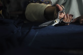 A hand holding a pair of metallic scissors is visible, poised to cut through a piece of dark fabric. The setting appears to be dimly lit, suggesting a focused, intimate workspace.