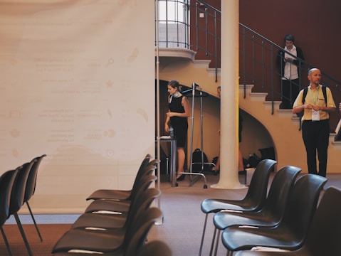 Several empty black chairs are arranged neatly in rows facing a large white projector screen or board. Behind the screen, a few people are standing and talking near a staircase in a venue that has a modern, professional ambiance. The stairway curves upward with railings, and the room has a warm color palette.