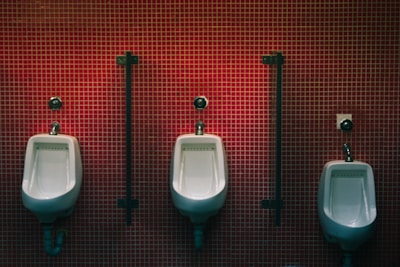 Three white urinals mounted on a tiled wall composed of small red tiles with dark vertical dividers between them.