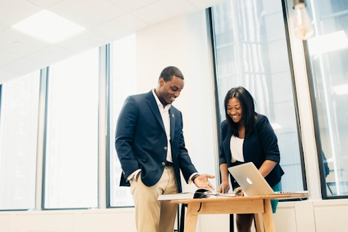 Two people are standing in an office space near a table with a laptop and papers. They are dressed in business attire and appear to be having a discussion.