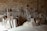 A small workshop with wooden boards and sawdust scattered on the floor. Various woodworking tools are mounted on the wall, including clamps and a saw. The brick wall in the background creates a rustic atmosphere, and there's a workbench filled with slabs of wood.