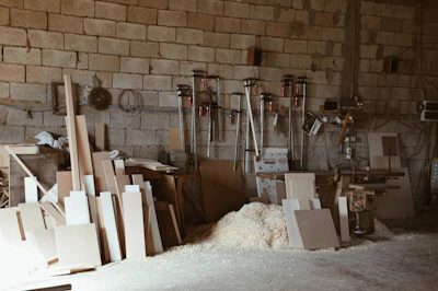 A small workshop with wooden boards and sawdust scattered on the floor. Various woodworking tools are mounted on the wall, including clamps and a saw. The brick wall in the background creates a rustic atmosphere, and there's a workbench filled with slabs of wood.