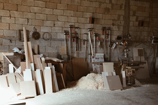 A small workshop with wooden boards and sawdust scattered on the floor. Various woodworking tools are mounted on the wall, including clamps and a saw. The brick wall in the background creates a rustic atmosphere, and there's a workbench filled with slabs of wood.