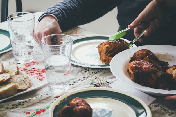 A table setting with plates of roasted chicken and a dish of sliced bread. Two glasses of water are placed on a table with a floral-patterned cloth. Hands are seen serving chicken onto the plates.