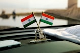 Two miniature Indian flags are positioned on a car dashboard with a small emblem between them. A smartphone lies nearby on the dashboard, and a cloth is draped over part of the surface. The background is blurred but suggests an outdoor setting.