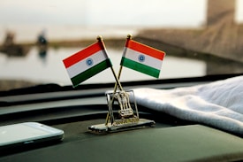 Two miniature Indian flags are positioned on a car dashboard with a small emblem between them. A smartphone lies nearby on the dashboard, and a cloth is draped over part of the surface. The background is blurred but suggests an outdoor setting.