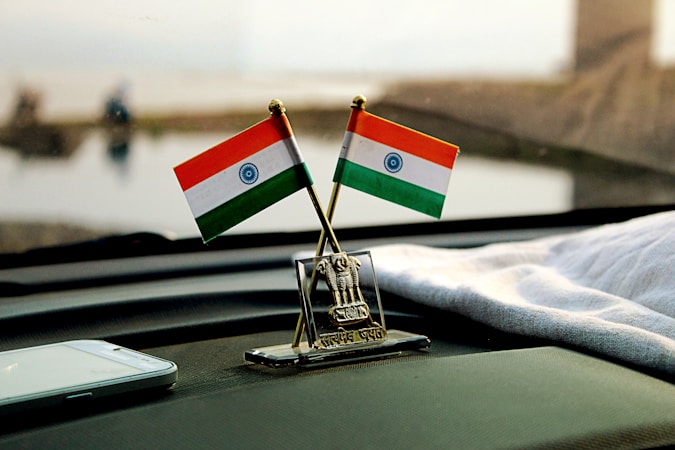 Two miniature Indian flags are positioned on a car dashboard with a small emblem between them. A smartphone lies nearby on the dashboard, and a cloth is draped over part of the surface. The background is blurred but suggests an outdoor setting.