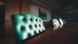 A long wooden conference table surrounded by tufted leather chairs. The chairs are upholstered in a deep green color, and the table is large with a polished finish. The room is dimly lit with ambient lighting from a window in the background, adding a warm glow. A painting hangs on the wall, partially visible in the soft lighting.
