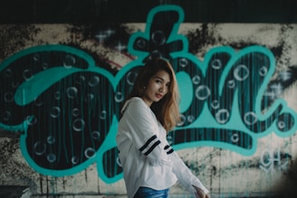 A young woman with long hair wearing a white shirt with black stripes on the sleeves is posed in front of a graffiti-covered wall. The graffiti features large, bold turquoise letters with circular patterns and drips, creating an urban and artistic backdrop.