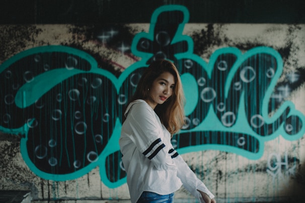 A young woman with long hair wearing a white shirt with black stripes on the sleeves is posed in front of a graffiti-covered wall. The graffiti features large, bold turquoise letters with circular patterns and drips, creating an urban and artistic backdrop.