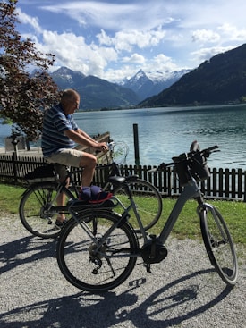 An elderly person rides a bicycle along a gravel path next to a serene lake surrounded by tall mountains. The scene features lush greenery, a wooden fence, and a tree with dark leaves. The sky is partly cloudy with patches of blue.