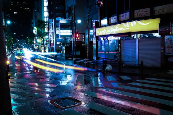 A nighttime urban scene featuring a wet street reflecting vibrant city lights. Neon signs illuminate the surroundings, with light trails from moving vehicles creating dynamic streaks across the road. The atmosphere is lively yet slightly mysterious.