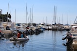 A marina filled with various boats and yachts lined up alongside each other. The water is calm, reflecting the vessels and the blue sky above. Several people are seen on a small inflatable boat navigating through the marina. The masts of the sailboats create vertical lines against the backdrop of a clear, sunny sky.