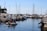 A marina filled with various boats and yachts lined up alongside each other. The water is calm, reflecting the vessels and the blue sky above. Several people are seen on a small inflatable boat navigating through the marina. The masts of the sailboats create vertical lines against the backdrop of a clear, sunny sky.