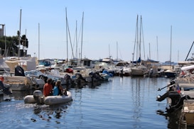 A marina filled with various boats and yachts lined up alongside each other. The water is calm, reflecting the vessels and the blue sky above. Several people are seen on a small inflatable boat navigating through the marina. The masts of the sailboats create vertical lines against the backdrop of a clear, sunny sky.
