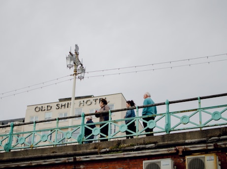 A street scene with a group of people walking across a bridge. The background features a building with the sign 'Old Ship Hotel.' There is a prominent streetlamp on the bridge, and strings of lights are visible against the cloudy sky.