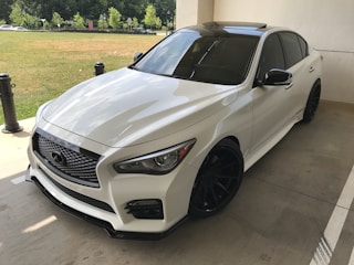 A sleek white sedan with tinted windows and black wheels parked on a concrete surface. The lighting accentuates the car's glossy finish and aerodynamic design. In the background, there are green trees and a grassy area, separated from the parking area by short black bollards.