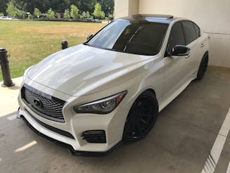 A sleek white sedan with tinted windows and black wheels parked on a concrete surface. The lighting accentuates the car's glossy finish and aerodynamic design. In the background, there are green trees and a grassy area, separated from the parking area by short black bollards.