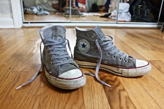 A pair of worn gray Converse high-top sneakers with white rubber soles and visible laces are placed on a wooden floor. Behind them, there is a reflective surface showing parts of a room with various objects.