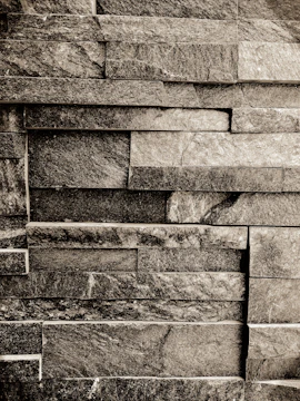 A textured wall made of rectangular stone slabs arranged in a staggered pattern. The stones vary in shade, creating a rustic and natural look.