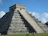 Smiling traveler standing in front of the towering pyramid at Chichen Itza under a bright blue sky.