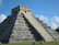 Smiling traveler standing in front of the towering pyramid at Chichen Itza under a bright blue sky.