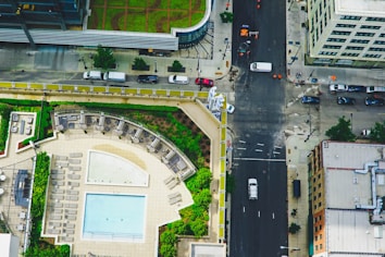 A rooftop with a swimming pool, surrounded by lounge chairs and green garden areas. The scene includes a city street below with cars parked along the sides and some vehicles moving. Buildings with varied architectural styles line the street.