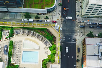 A rooftop with a swimming pool, surrounded by lounge chairs and green garden areas. The scene includes a city street below with cars parked along the sides and some vehicles moving. Buildings with varied architectural styles line the street.