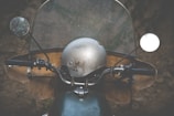 A close-up view of a vintage motorcycle with a metal helmet resting on it. The motorcycle features a large transparent windshield with two round mirrors protruding from either side. The background is blurred, focusing attention on the motorcycle's handlebars and helmet.