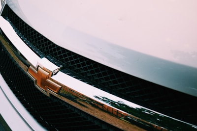 A close-up of a car grille featuring a prominent emblem of a gold bowtie set against a chrome and black lattice background with reflections of the surrounding environment.