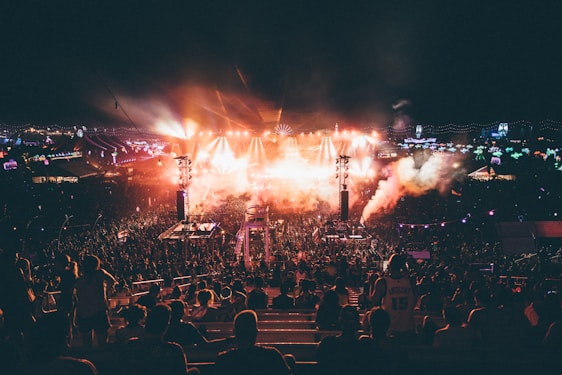 A large outdoor music festival during the night featuring a crowd of people gathered around a well-lit stage. Bright lights and smoke effects add to the energetic atmosphere. The scene is dynamic with colorful lights and vibrant overhead decorations.