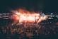 A large outdoor music festival during the night featuring a crowd of people gathered around a well-lit stage. Bright lights and smoke effects add to the energetic atmosphere. The scene is dynamic with colorful lights and vibrant overhead decorations.