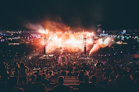 A large outdoor music festival during the night featuring a crowd of people gathered around a well-lit stage. Bright lights and smoke effects add to the energetic atmosphere. The scene is dynamic with colorful lights and vibrant overhead decorations.