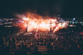 A large outdoor music festival during the night featuring a crowd of people gathered around a well-lit stage. Bright lights and smoke effects add to the energetic atmosphere. The scene is dynamic with colorful lights and vibrant overhead decorations.