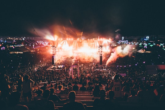 A large outdoor music festival during the night featuring a crowd of people gathered around a well-lit stage. Bright lights and smoke effects add to the energetic atmosphere. The scene is dynamic with colorful lights and vibrant overhead decorations.