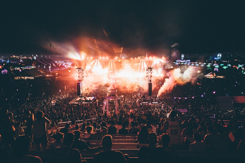 A large outdoor music festival during the night featuring a crowd of people gathered around a well-lit stage. Bright lights and smoke effects add to the energetic atmosphere. The scene is dynamic with colorful lights and vibrant overhead decorations.