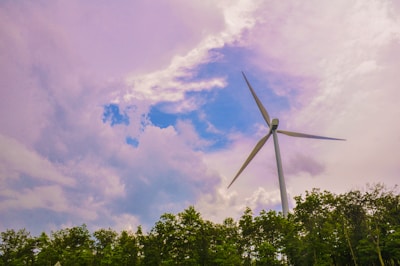 A large wind turbine towers over a dense row of green trees. The sky is filled with dramatic clouds, ranging from white and fluffy to darker purple tones, with patches of clear blue sky peeking through.